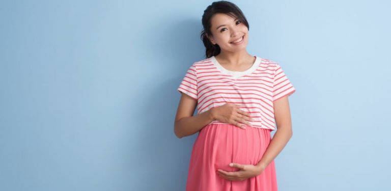 A pregnant woman holding her stomach, standing against a blue wall.