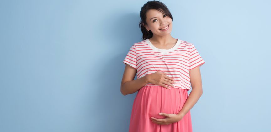 A pregnant woman holding her stomach, standing against a blue wall.