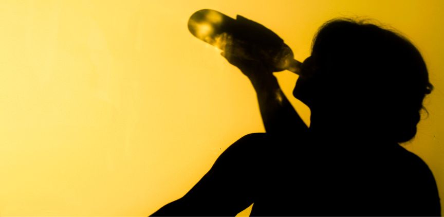 A silhouette of a woman drinking from a bottle of alcohol against a yellow background.