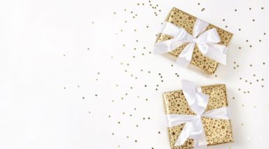 Two presents wrapped in gold paper with white ribbon, on a white background with gold glitter scattered around.