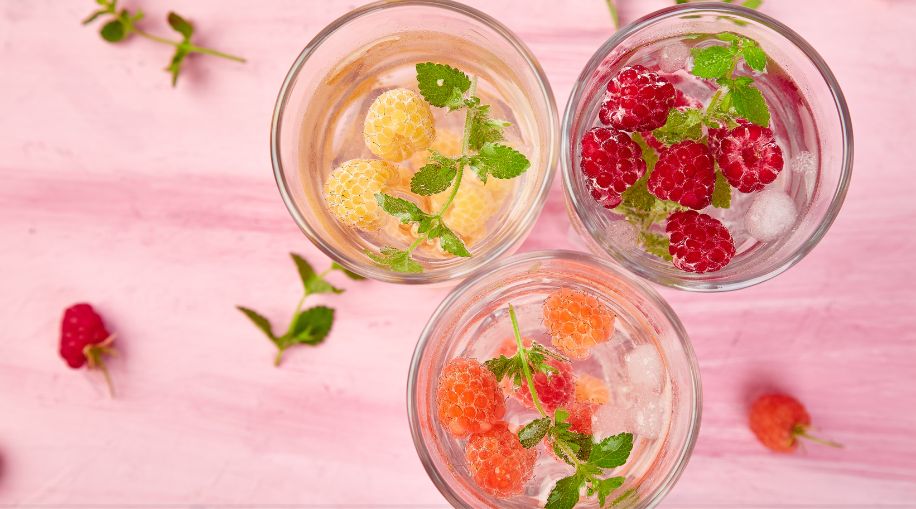 A top-down view of three glasses with raspberries in them, on a pink background.